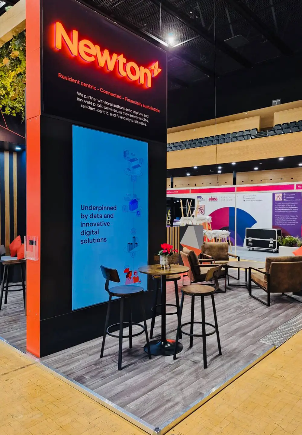 Modern exhibitor booth with Newton sign, digital display, tall stools, and lounge seating with tables.