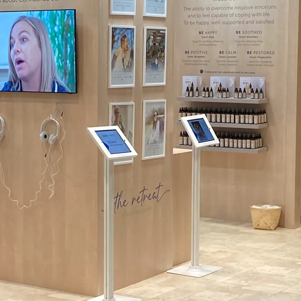 Retail display area with wall-mounted video, headphones, product shelves, framed photos, and touchscreen stands.