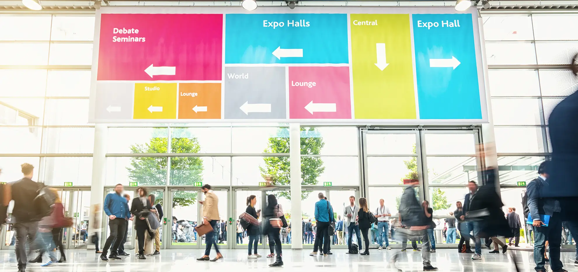 People walking and standing under a colorful overhead sign with directions in a large bright expo hall.