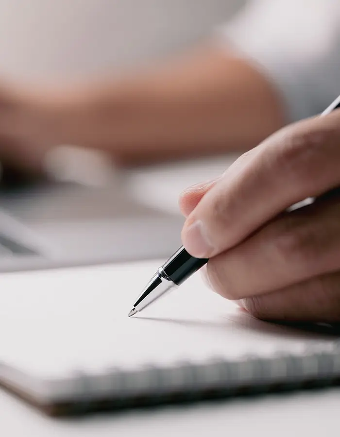 Hand holding a black pen poised to write on a blank white notebook page.