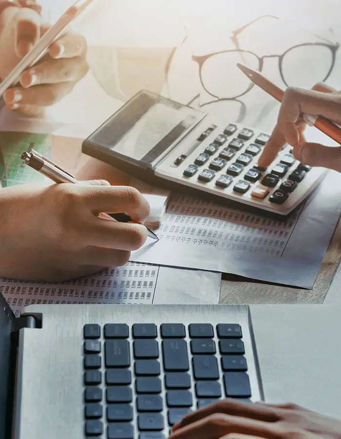 Two people working with calculator, pen, paper with numbers, and laptop keyboard on a desk.