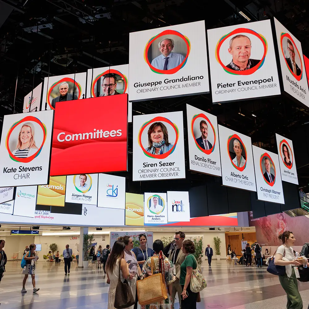 People gather under a ceiling display showing committee member portraits and names at a conference venue.