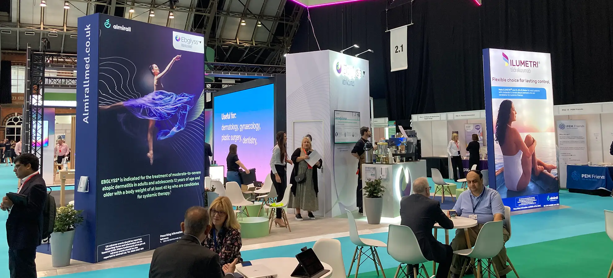 Exhibition booth with banners for Almirall featuring Ebgalyss and Ilumetri products with people talking and sitting.