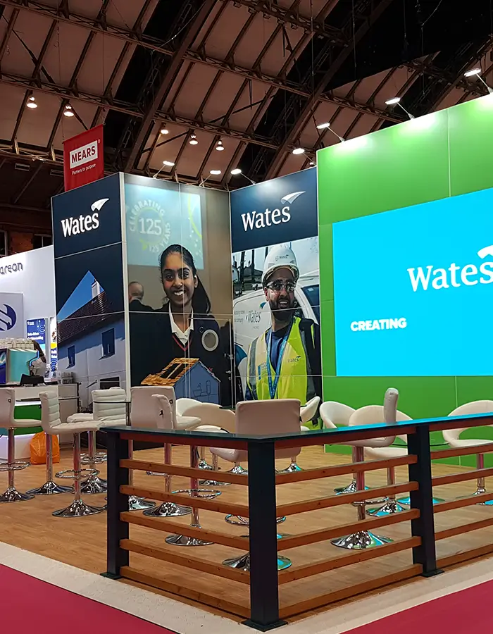 Wates company booth at event with chairs, large video screen, and photos of workers in safety gear.