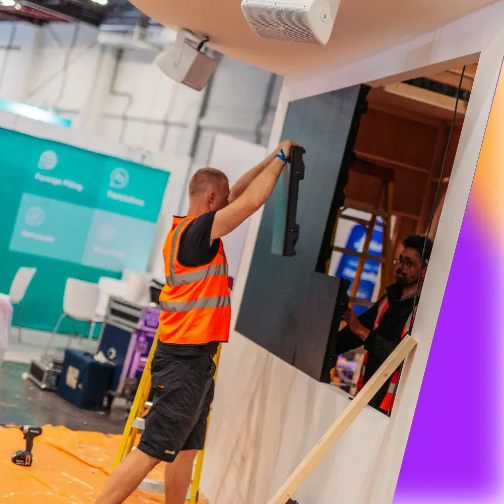 Worker in an orange safety vest installing a large screen or panel on a structure indoors.