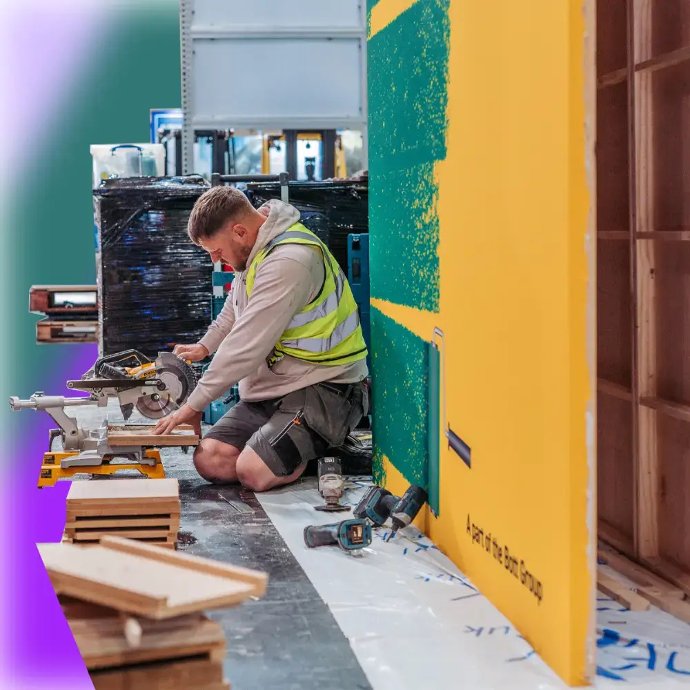 Worker in a yellow safety vest kneels using a miter saw to cut wood pieces indoors.