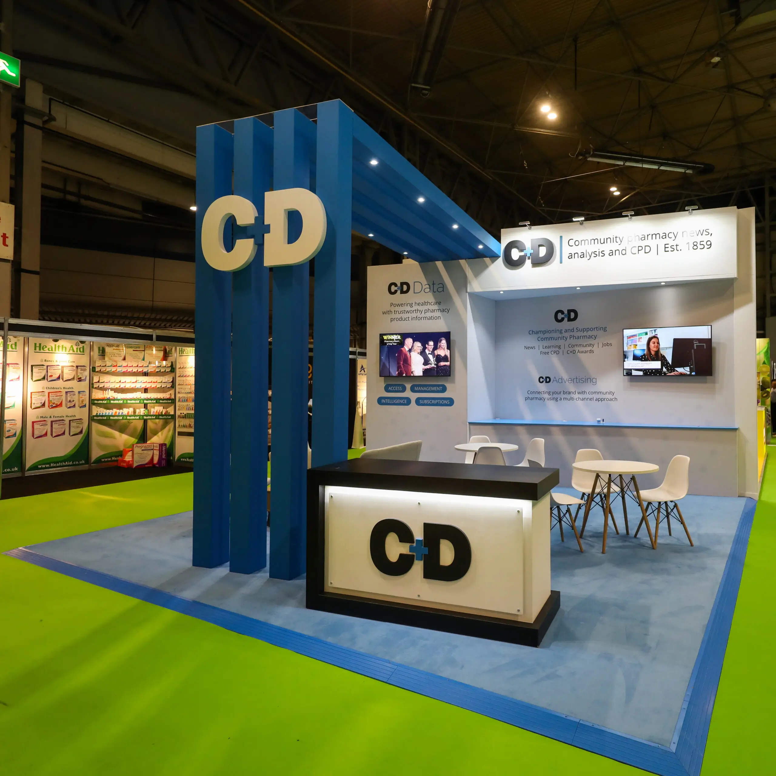 Exhibition booth with large C+D logo, blue pillars, seating area, and promotional screens in a hall.