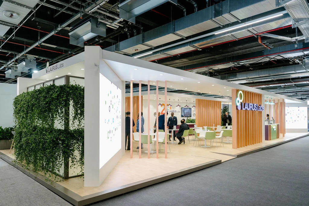 Modern exhibition booth for Laurus Labs with seating, green plants, and people conversing inside.
