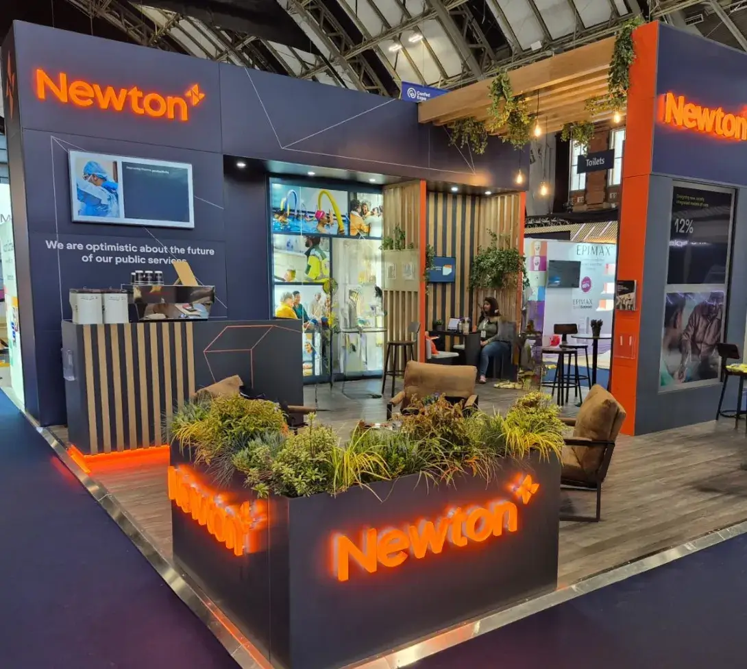 Newton branded exhibition booth with seating, plants, digital screens, and a person seated inside.
