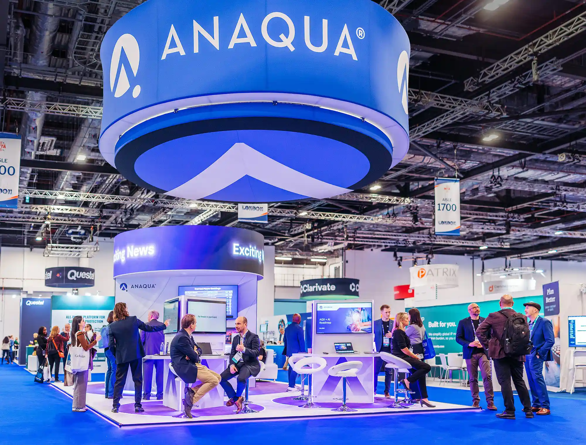 People interacting at ANAQUA booth with bright blue displays and large overhead ANAQUA logo at a conference.