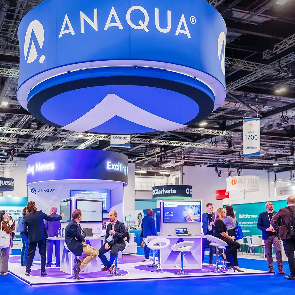 Trade show booth with ANAQUA branding and people talking and viewing screens under a large hanging sign.