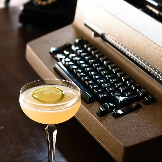 Cocktail in a coupe glass with a lime wheel garnish next to a vintage typewriter on a dark wood surface