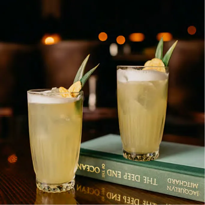 Two tall cocktails with pineapple garnish on a stack of books, with warm bar lights in the background