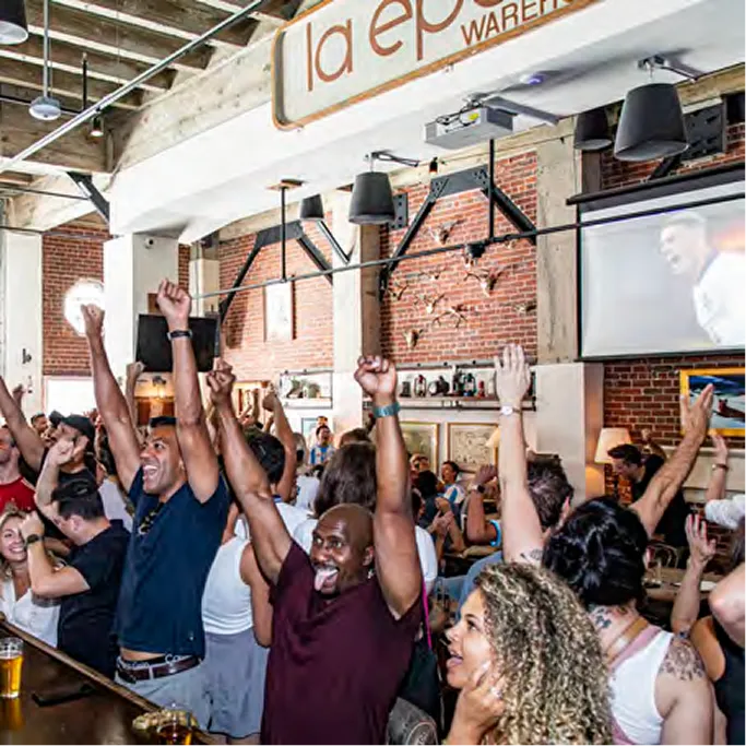 Excited crowd with arms raised watching sports at La Epoca Warehouse bar with exposed brick walls