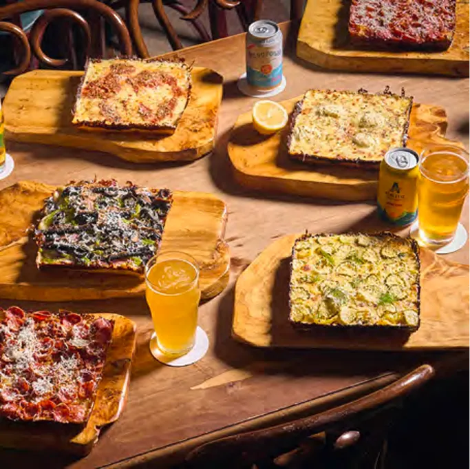 Overhead spread of Detroit-style square pizzas on wooden boards with cans and glasses of beer on a wooden table