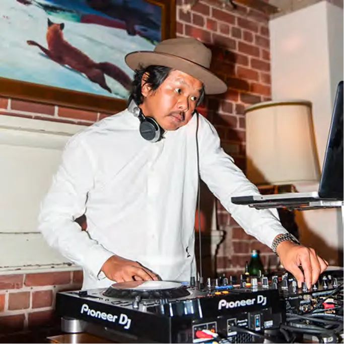 DJ performing at Pioneer DJ turntables wearing a wide-brim hat and white shirt, with brick walls and a fox painting behind