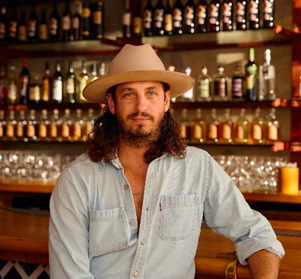 Man with long curly hair and beard wearing a wide-brim hat, leaning on a bar with a wall of spirits behind him