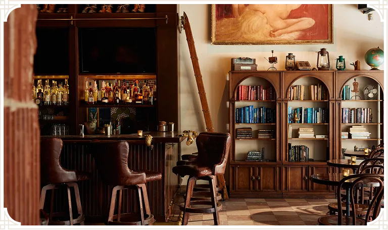Lost Boy bar corner with leather bar stools, backbar spirits, and a vintage arched bookshelf with books and antique decor