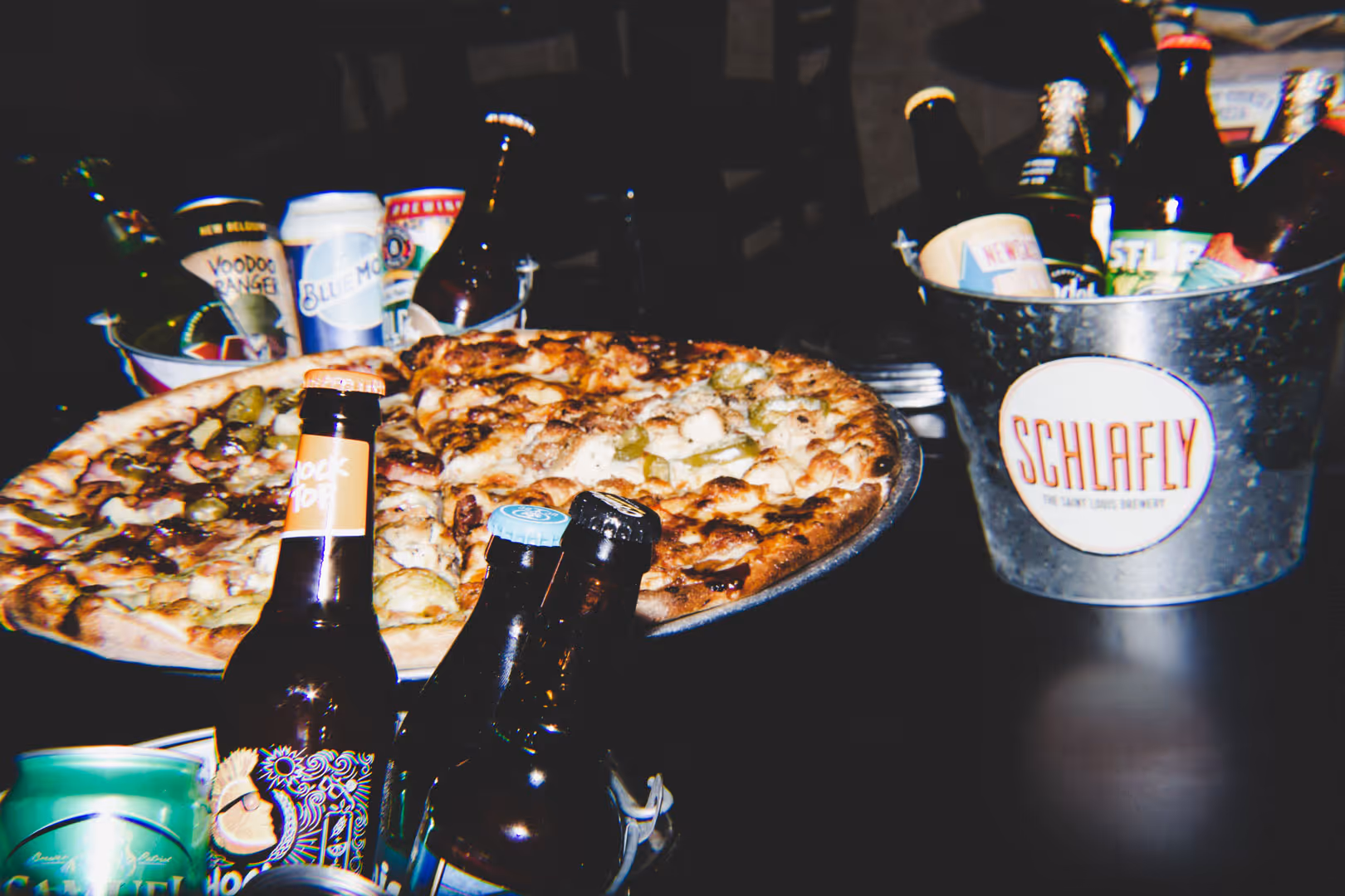 Large pizza on metal tray surrounded by various beer bottles and cans in a metal bucket.