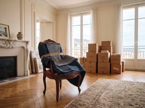 Un intérieur d'appartement lumineux à Cannes avec un fauteuil ancien soigneusement protégé par une couverture de déménagement près de cartons bien rangés.