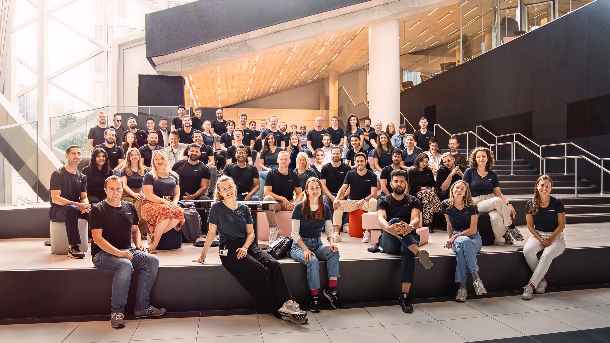 Ein großes Teamfoto von Leuten, die in einem modernen Gebäude mit Treppen und Glas sitzen und lächeln.