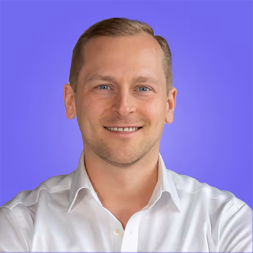 Ein lächelnder Mann mit hellbraunen Haaren, der ein weißes Hemd trägt, vor einem violetten Hintergrund.