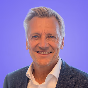 Porträt eines lächelnden älteren Mannes mit grauen Haaren, der ein weißes Hemd und einen dunkelblauen Blazer trägt, vor einem violetten Hintergrund.