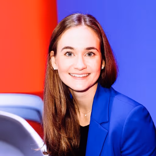 Smiling young woman with long brown hair wearing a blue blazer and black top, posed against a red and blue background.