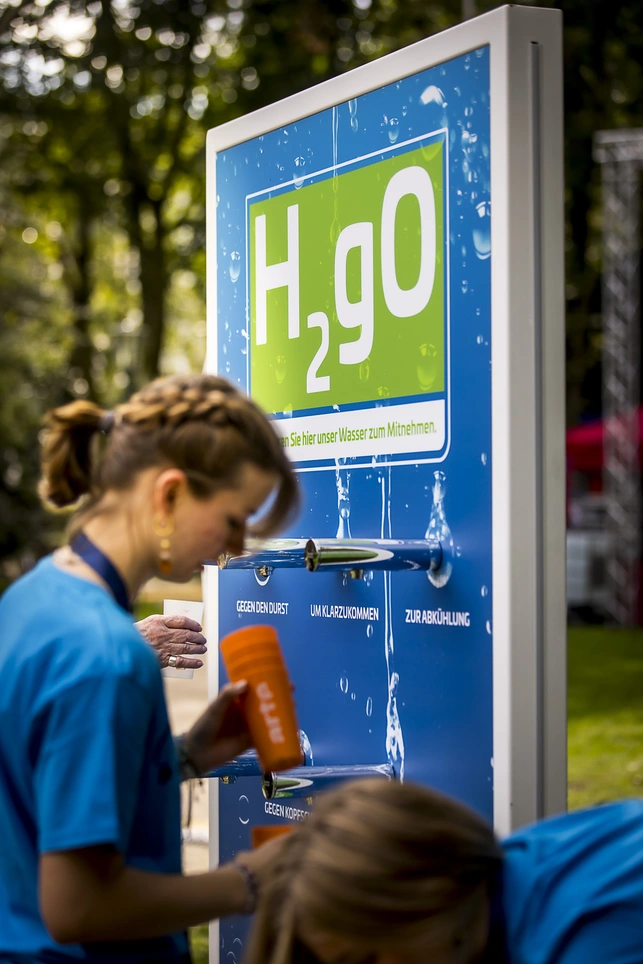 Wasserspender mit H2Go-Logo und zwei Personen füllen eine orangefarbene Flasche im Park.