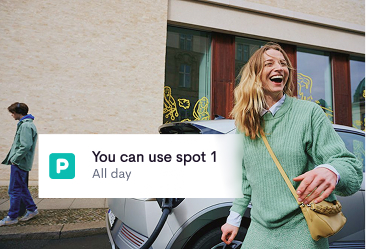 A smiling woman in a green sweater holding a small bag stands next to a parked gray car near a building with large windows, accompanied by an on-screen parking notification saying, 'You can use spot 1 All day.'