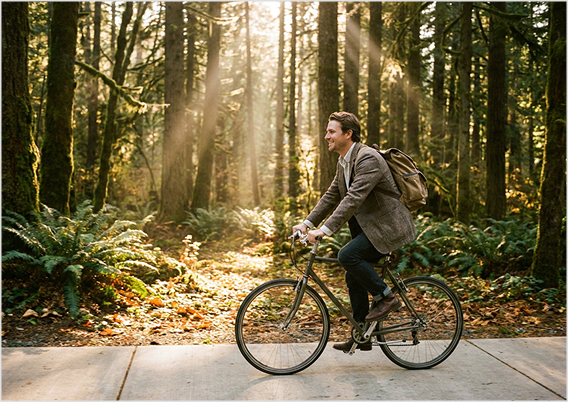 A man wearing a brown blazer and backpack rides a bicycle on a paved path through a sunlit forest with tall trees and green ferns.