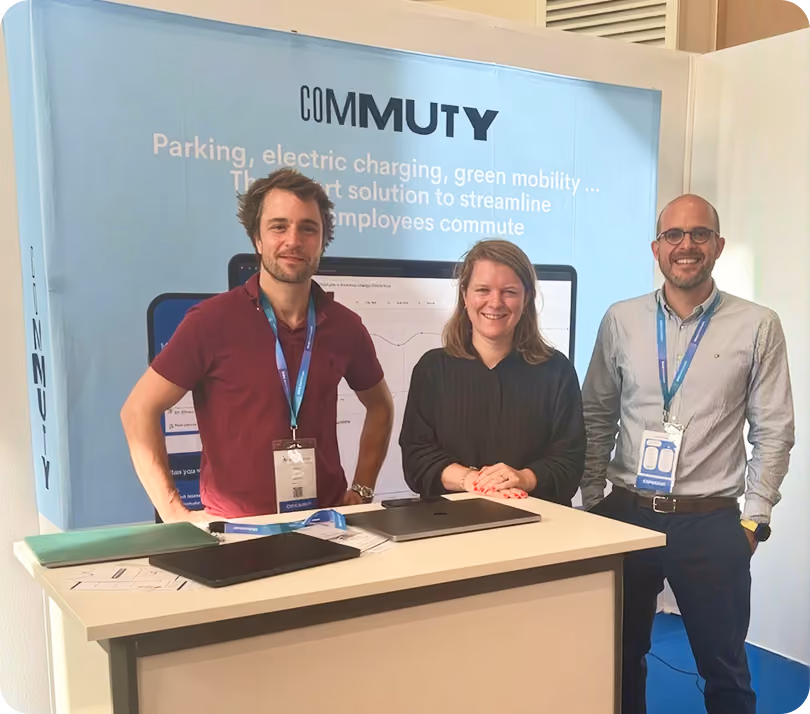 Three smiling people, two men and a woman, standing behind a white booth table with laptops and documents, in front of a blue backdrop displaying the word 'COMMUTY' and text about parking, electric charging, and green mobility solutions.
