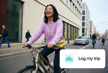 A smiling woman in a purple sweater rides a bicycle on a city street with buildings and other cyclists in the background.