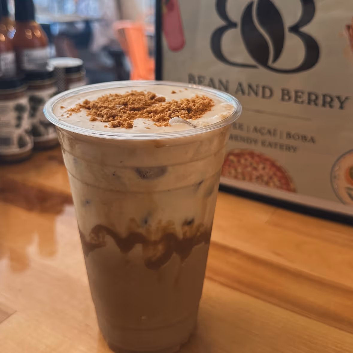 Iced coffee drink topped with cream and brown crumbles in a plastic cup on a wooden table.