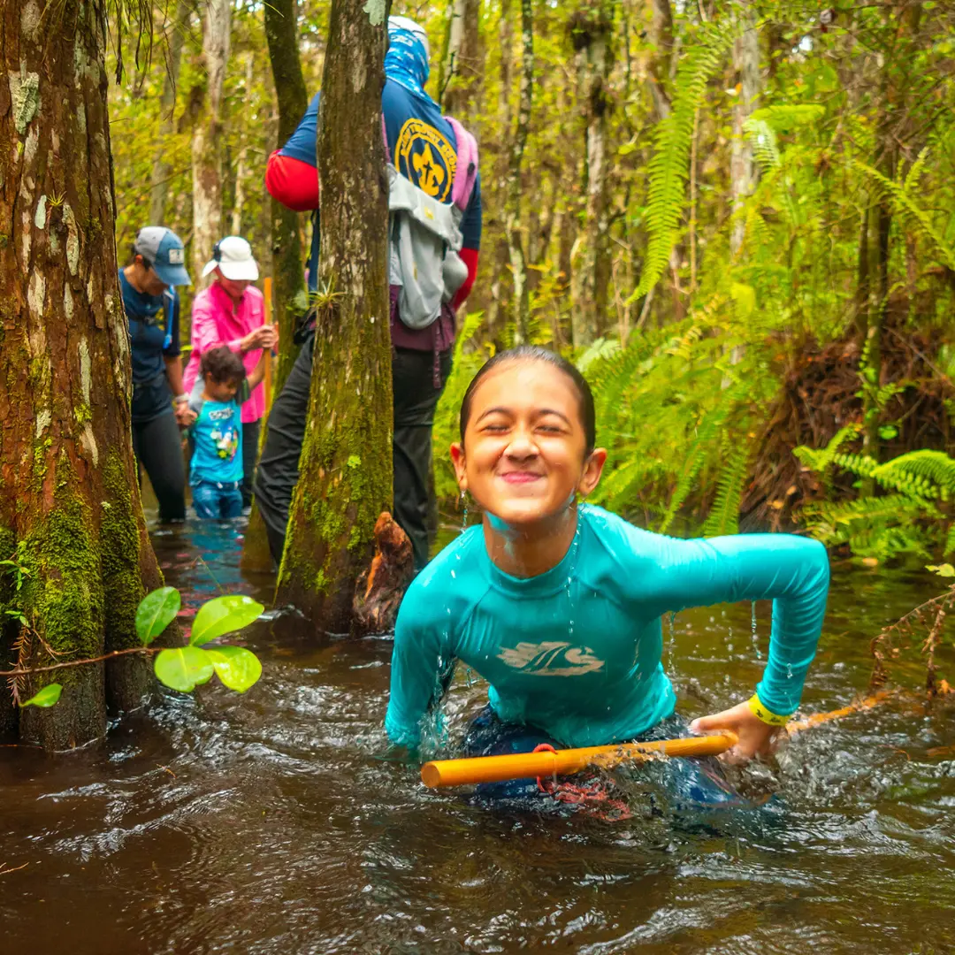 Child smiling and squinting while wading in forest water holding a yellow stick, with adults and kids in background.