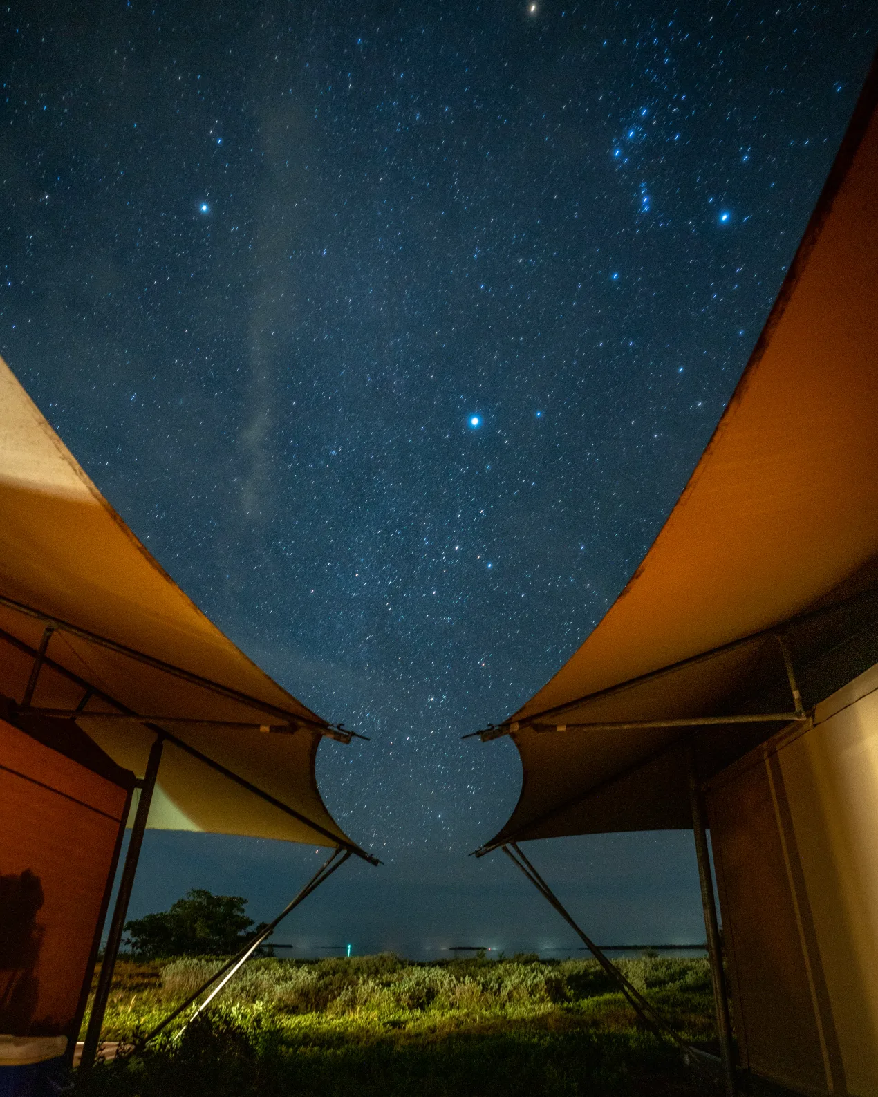 Starry night sky viewed between two angular tent canopies over grassy terrain.