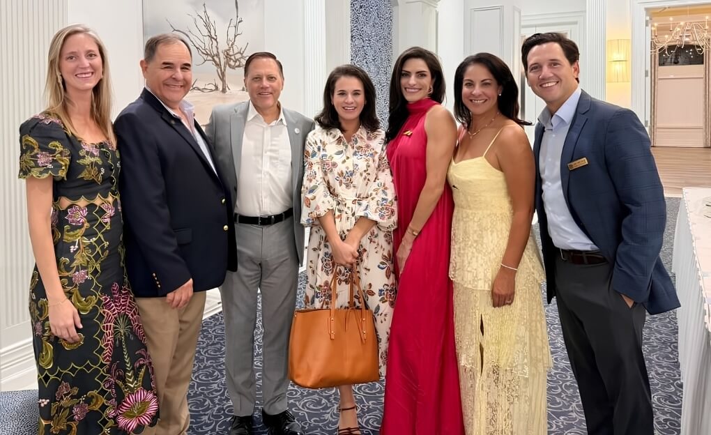 Group of seven people dressed formally standing side by side indoors smiling at the camera.