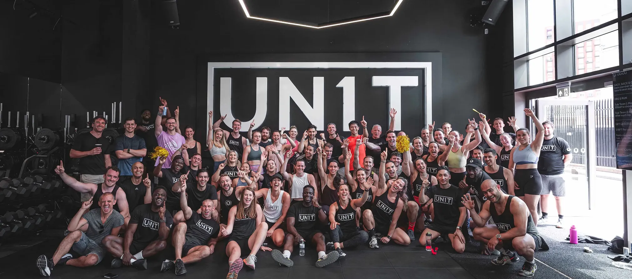Group of people in a gym stretch arms down and up in front of a UN1T sign and dumbbell rack.