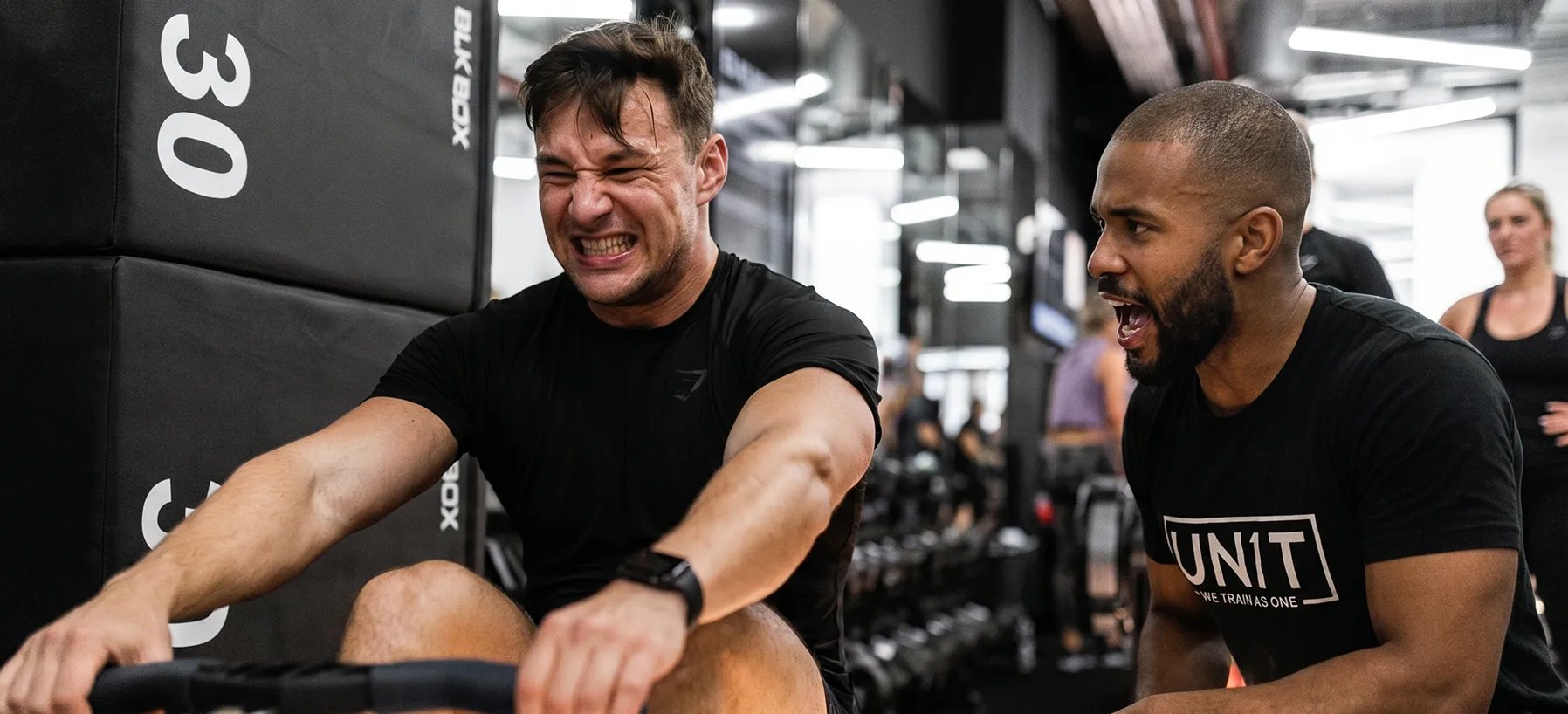 Man grimacing while working out on rowing machine, trainer encouraging him nearby in gym.