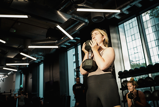 Woman holding a kettlebell in a gym with other people and large windows in the background.