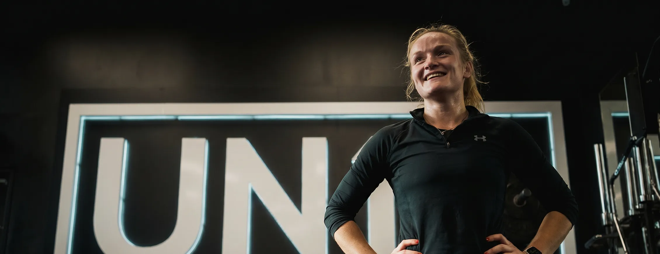 Smiling woman in black athletic shirt standing with hands on hips in a gym with neon sign behind her.