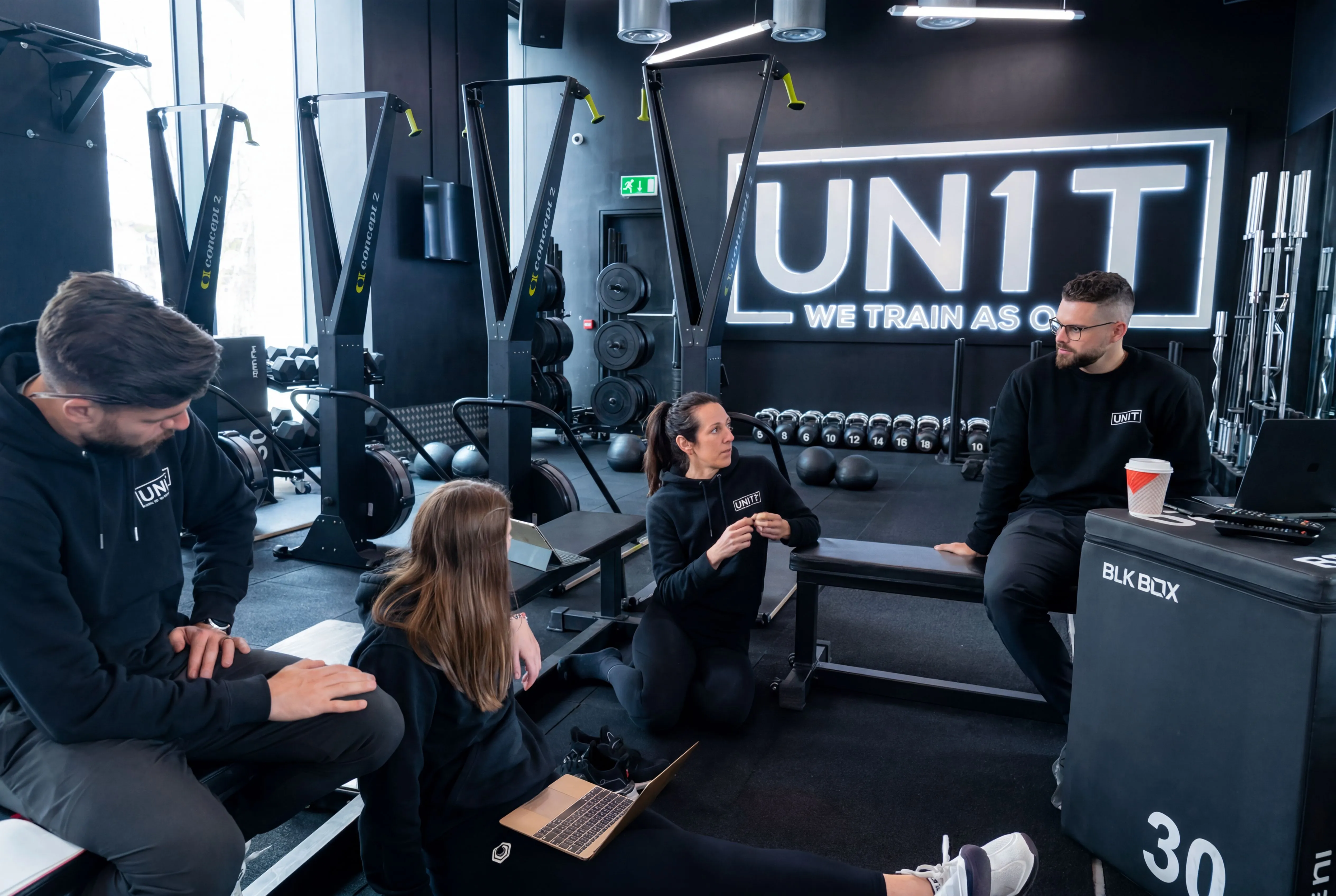 Fitness coach speaking to a line of adults standing in a gym with rowing machines and black walls.