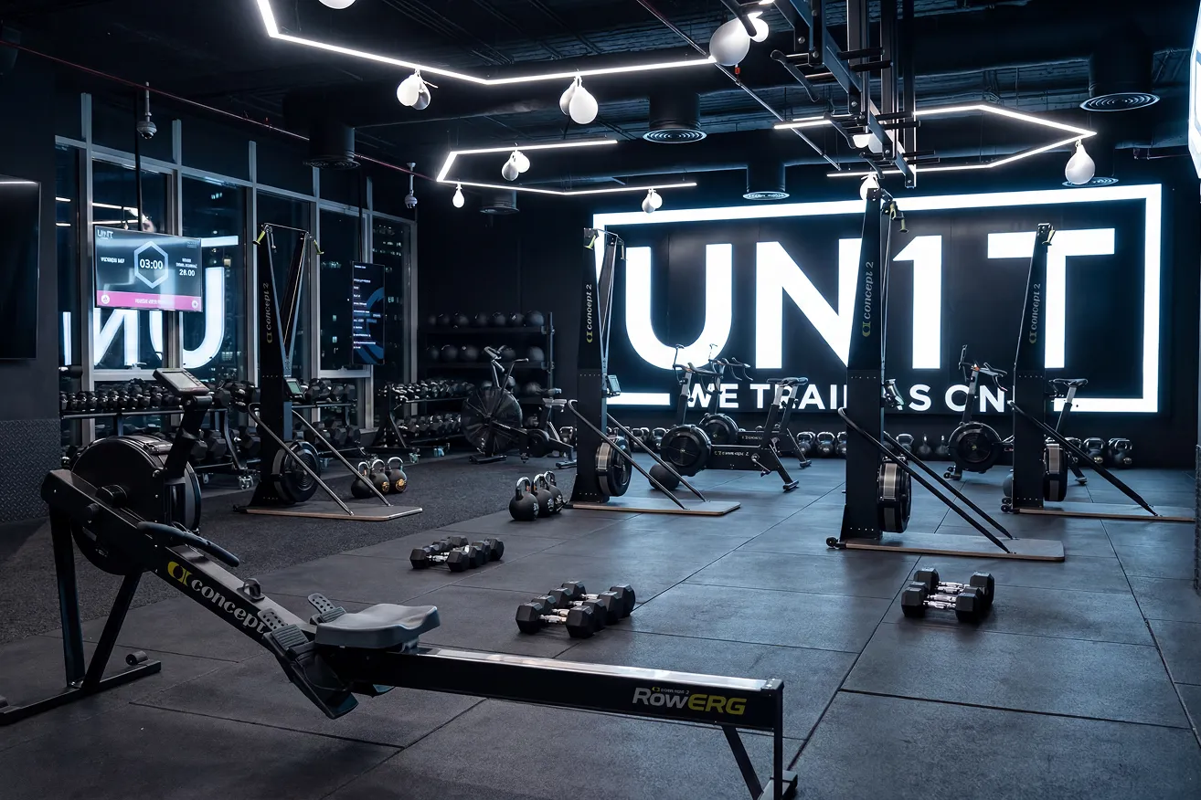 Modern indoor gym with rowing machines, weights, kettlebells, and large illuminated UN1T sign.