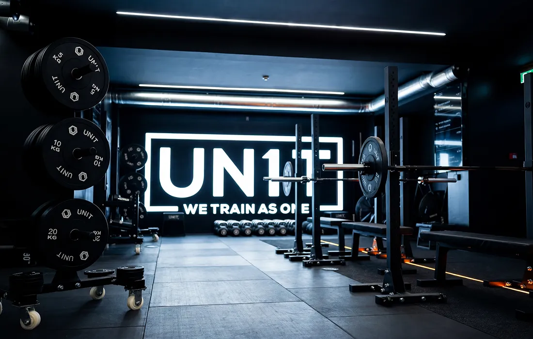 Modern gym interior with weights, barbells, benches under ceiling lights and UNIT logo on back wall.