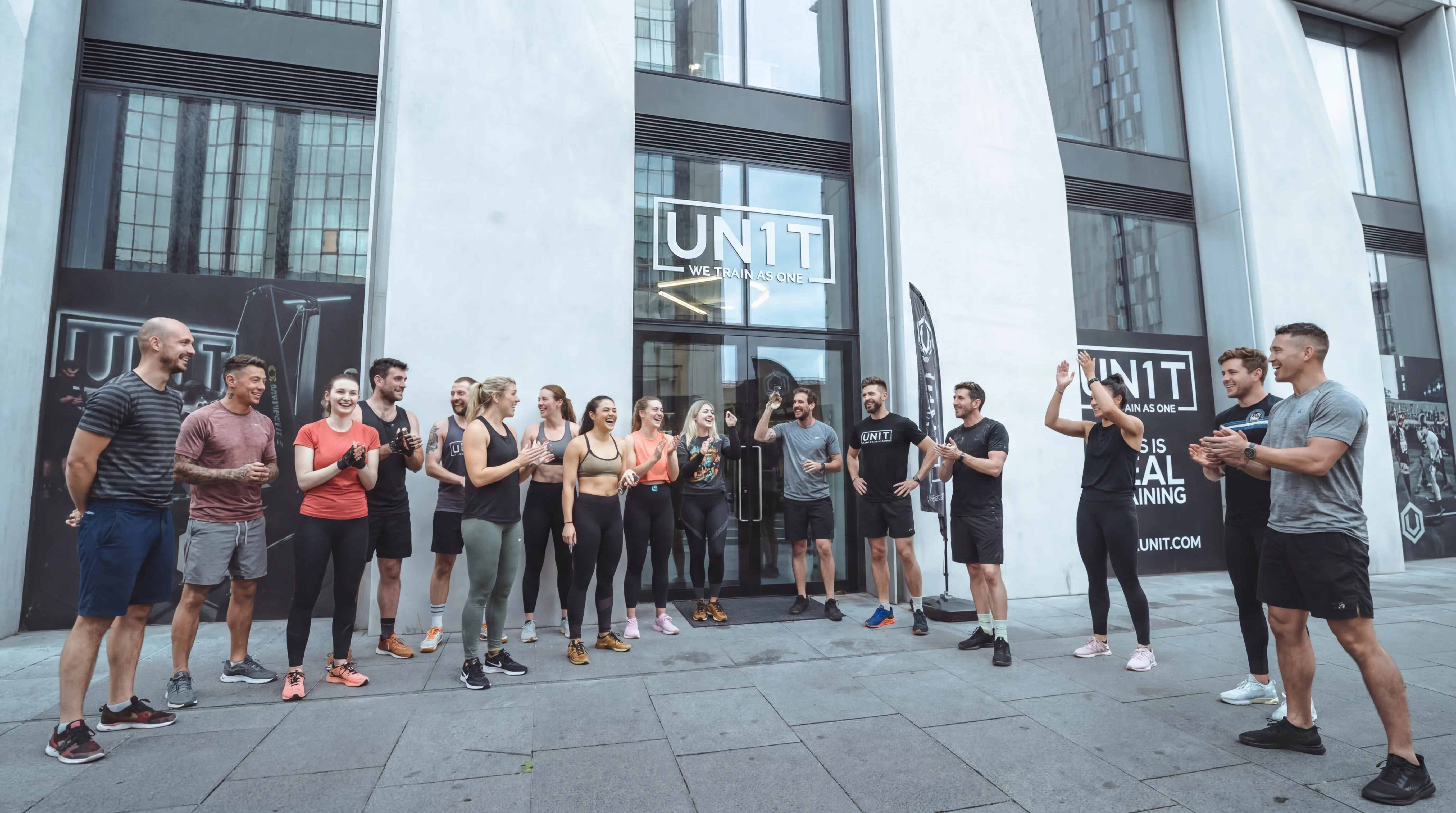 Group of people in athletic wear standing outside UNIT gym, cheering and clapping.