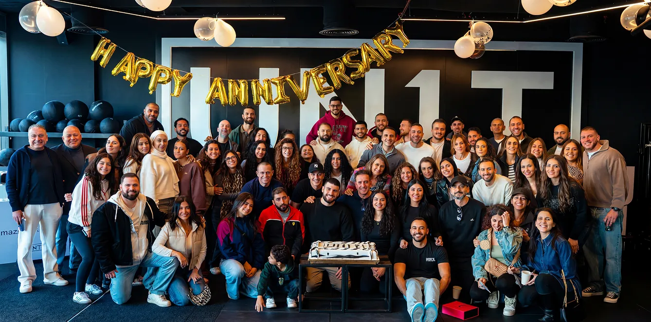 Large group of people smiling under gold balloons spelling Happy Anniversary with a cake in front.