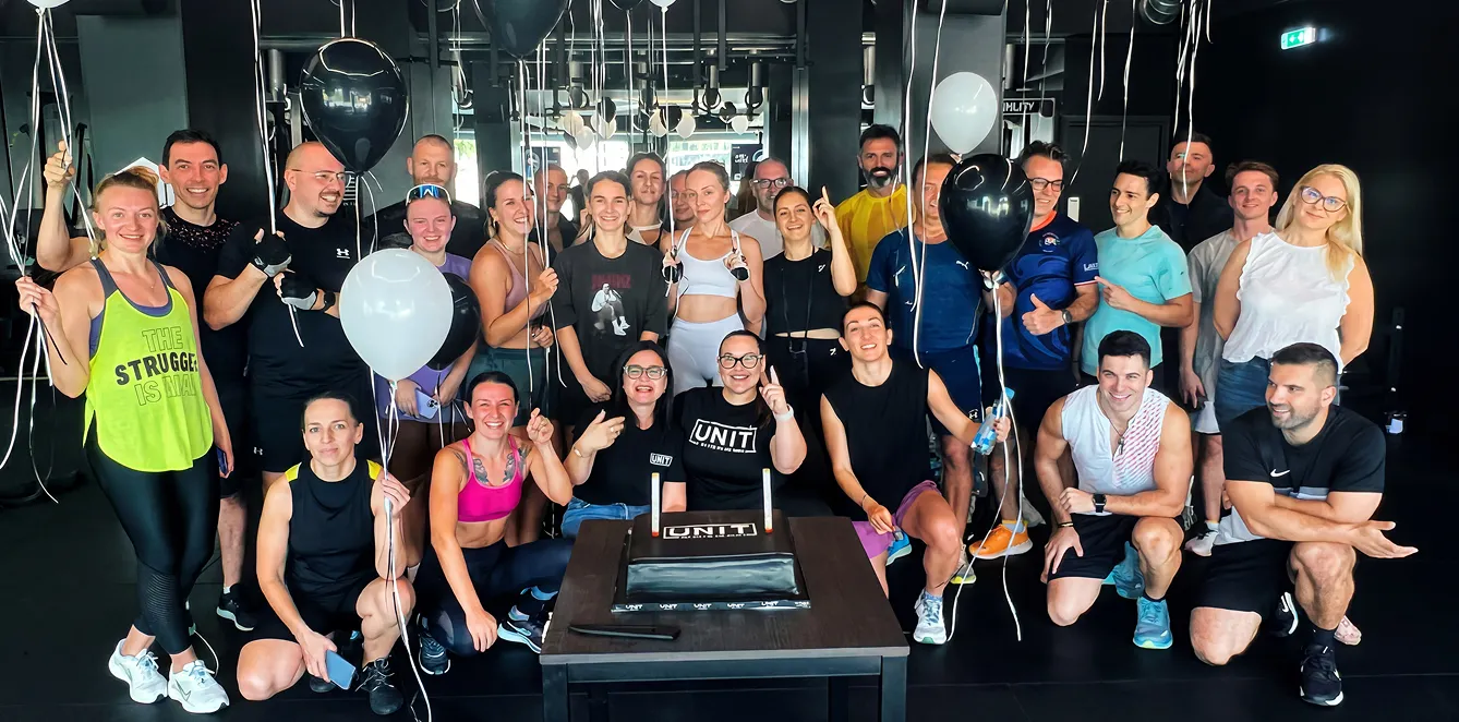 Group of fit people posing with balloons around a table with a UNIT-branded cake in a gym.