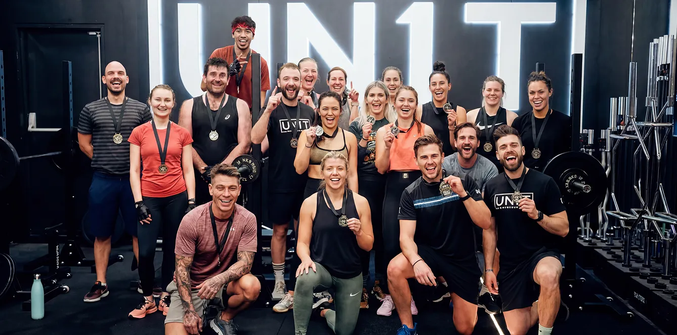 Group of people in workout clothes smiling and showing medals in a gym with 'UNIT' sign behind them.