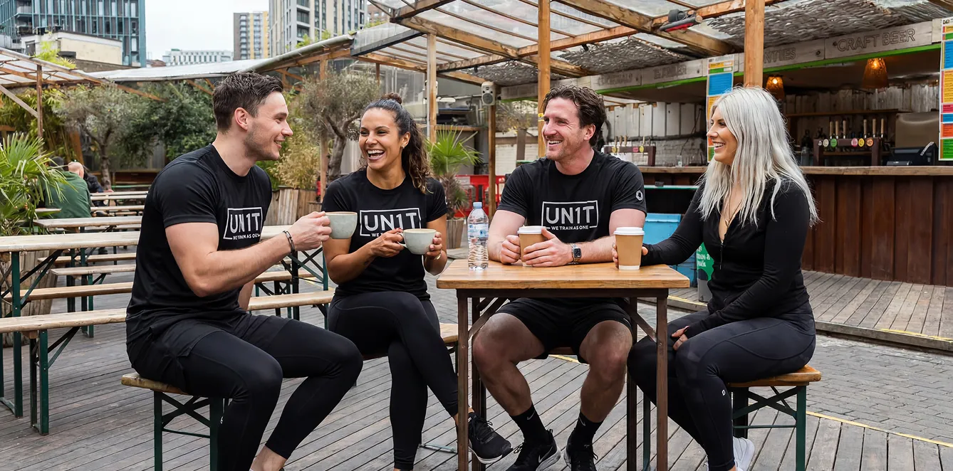 Four UN1T couaches in black workout clothes sit outside at a wooden table laughing and holding coffee cups.
