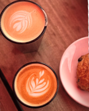 Two cups of latte with leaf latte art on a wooden table next to a plate with pastry.